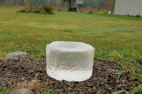 A large, clear ice block sits on a rocky surface in a grassy field, surrounded by trees and a distant building.