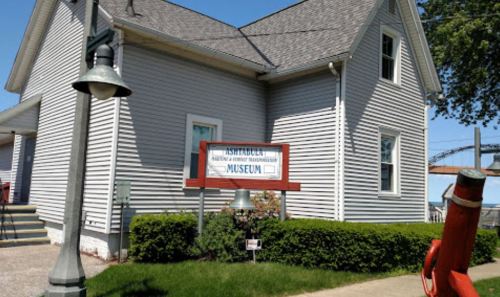 A gray house with a sign reading "Ashtabula Maritime and History Museum" in front, surrounded by greenery.