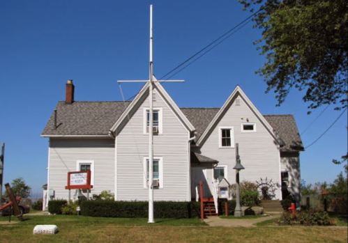 A white house with a steep roof, flagpole, and garden, set against a clear blue sky.