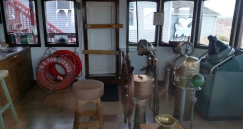 A nautical-themed room with a ship's wheel, vintage equipment, and a life preserver, featuring large windows.