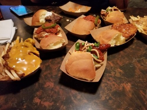 A variety of sliders and fries served in paper trays on a table, featuring different toppings and sauces.
