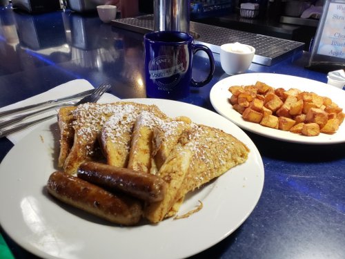 A plate of French toast dusted with powdered sugar, sausage links, and a side of golden-brown home fries.