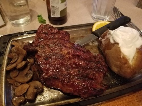 A sizzling steak with mushrooms, a baked potato topped with sour cream, and a lemon slice on a metal plate.