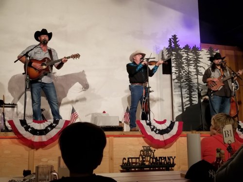 A country band performs on stage with guitars and a violin, decorated with patriotic bunting and a horse mural in the background.