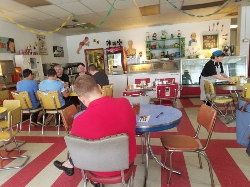 A retro diner with colorful chairs, patrons seated at tables, and a counter displaying vintage decor and memorabilia.