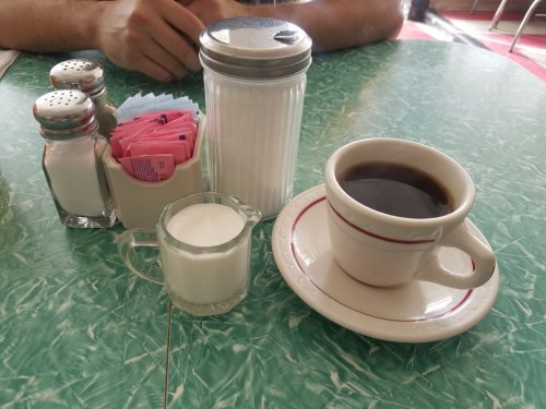 A cup of black coffee on a saucer, with a small pitcher of milk and sugar packets on a green table.