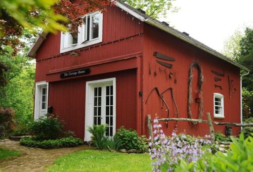 A charming red cottage with white windows, surrounded by greenery and flowers, featuring rustic tools on the wall.