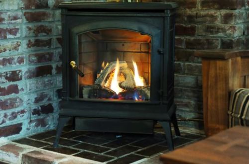 A cozy fireplace with flames burning inside, set against a brick wall and wooden furniture nearby.