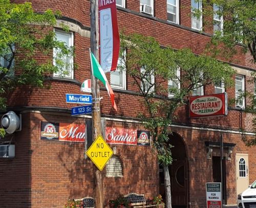 A brick building with a pizza restaurant sign, Italian flag, and street signs at the corner of E 123 St and Mayfield.