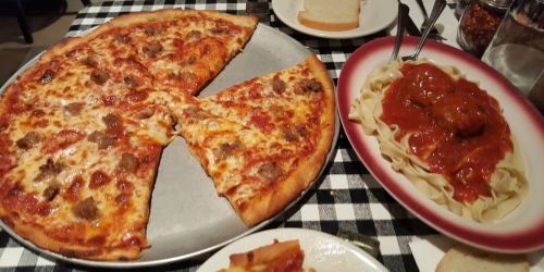A large pizza with slices cut out, alongside a plate of spaghetti and meatballs on a checkered tablecloth.