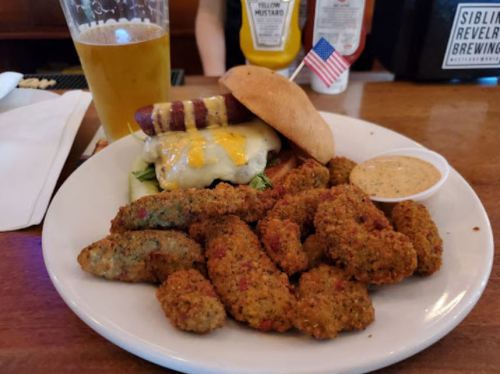 A plate with a cheeseburger topped with a hot dog, fried pickles, and a dipping sauce, alongside a glass of beer.