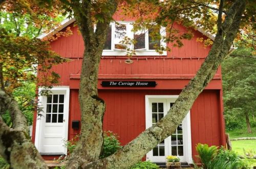 A red wooden building with a sign reading "The Carriage House," framed by a tree and surrounded by greenery.