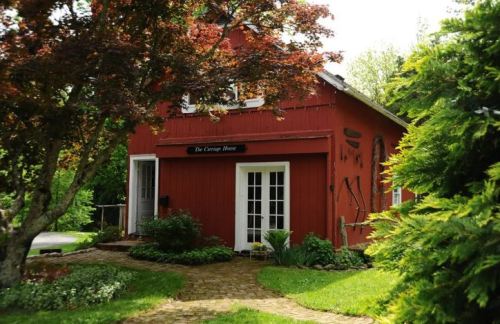 A charming red cottage surrounded by greenery and a stone pathway, featuring a sign that reads "The Carriage House."