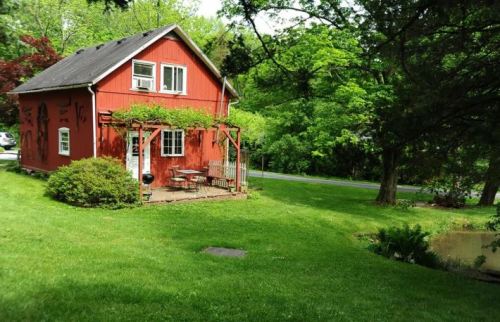 A charming red house with a porch, surrounded by lush green grass and trees, near a small pond.