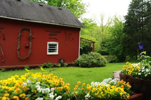 A red barn surrounded by green grass and colorful flower beds, with trees in the background.