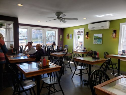 A cozy restaurant interior with wooden tables, black chairs, and colorful walls, featuring large windows and artwork.