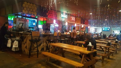A lively bar with colorful lights, wooden tables, and a bustling atmosphere, featuring a long counter and patrons enjoying drinks.