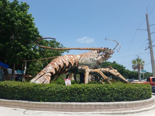 Giant lobster sculpture surrounded by greenery, set against a clear blue sky and power lines in the background.