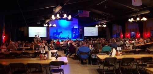 A large indoor venue filled with people seated at tables, with a stage and screens in the background, decorated for an event.