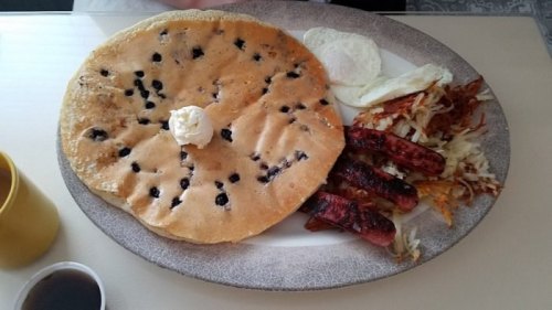 A large blueberry pancake with butter, two eggs, hash browns, and sausage links on a plate.
