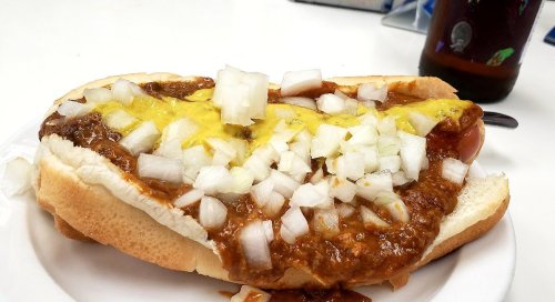 A hot dog topped with chili, mustard, and diced onions on a white plate. A drink is partially visible in the background.
