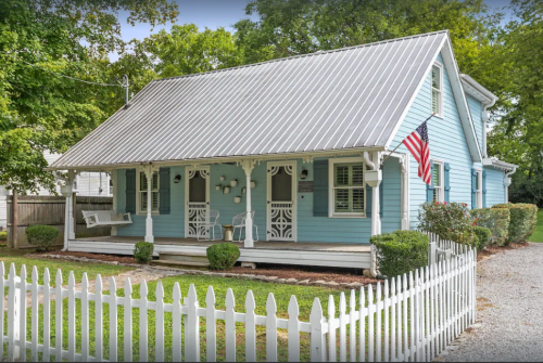 A charming blue house with a metal roof, white picket fence, and American flag, surrounded by greenery.