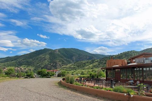 Scenic view of mountains and a rustic building with outdoor seating under a blue sky with fluffy clouds.