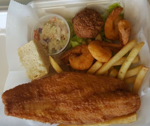 A takeout container with fried fish, shrimp, coleslaw, fries, a roll, and a fried chicken piece on a bed of lettuce.