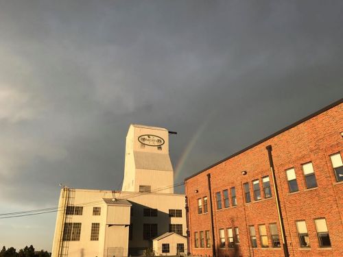 A tall industrial building with a sign, under a cloudy sky with a faint rainbow in the background.