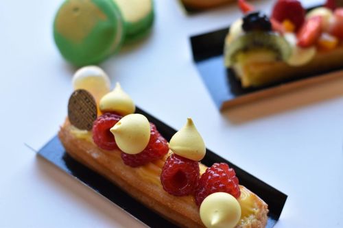 A close-up of a pastry topped with yellow cream, raspberries, and decorative elements, with macarons in the background.