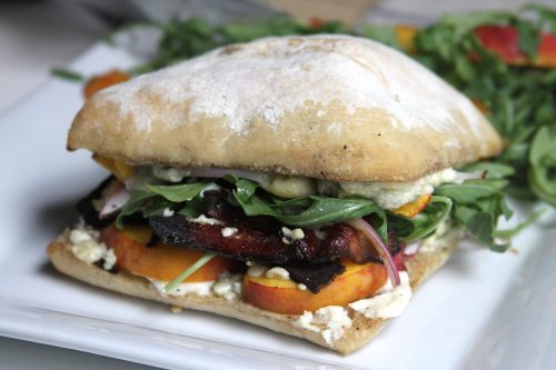 A delicious sandwich with layers of greens, peaches, and cheese on a crusty bread, served on a white plate.