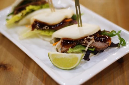 Three bao buns filled with meat, vegetables, and sauce, garnished with a lime wedge on a white plate.