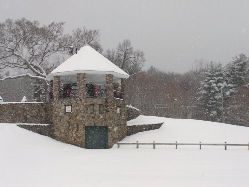 A stone gazebo covered in snow, surrounded by a winter landscape with falling snowflakes and trees in the background.