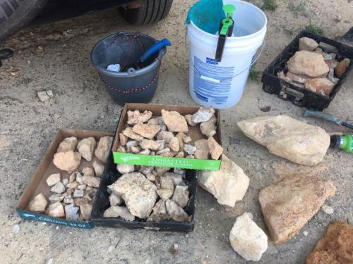 Three boxes of various rocks on the ground, with a bucket and a container nearby.