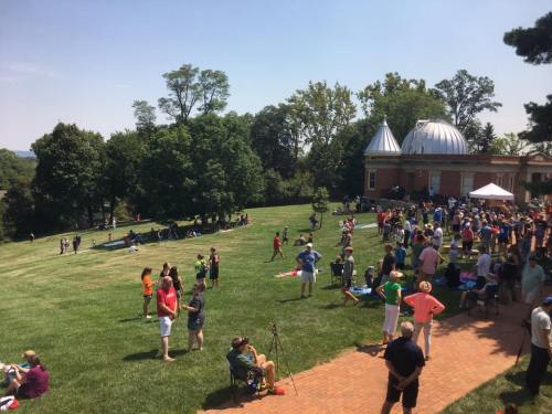 A large crowd gathers on a sunny day in a grassy area near a building with domed roofs, enjoying outdoor activities.