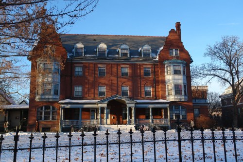 A large, historic brick building with a steep roof, surrounded by snow and a wrought iron fence on a clear day.