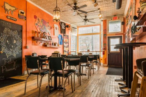 Cozy café interior with orange walls, wooden floors, and empty chairs, featuring colorful artwork and a sunny window.