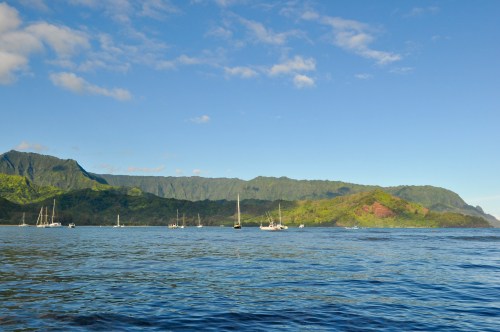 A serene coastal view with sailboats on calm waters, surrounded by lush green mountains under a clear blue sky.