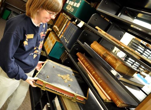 A woman in a library smiles while handling a large, ornate book on a shelf filled with various books.