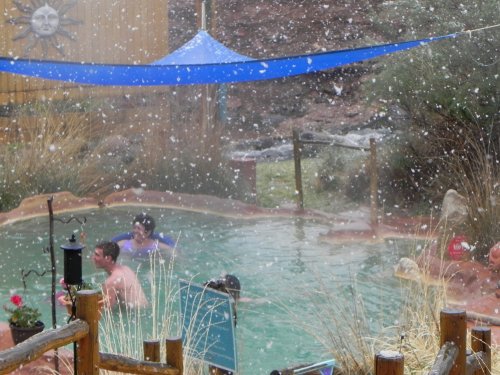 People enjoying a hot spring pool while snow falls around them, with a blue tarp overhead and a scenic backdrop.