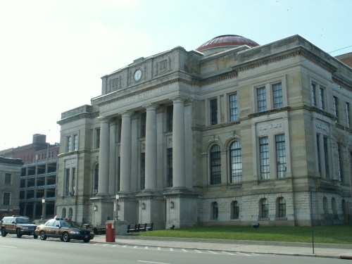Historic building with a clock, large columns, and a red dome, set against a clear sky. Cars parked in front.