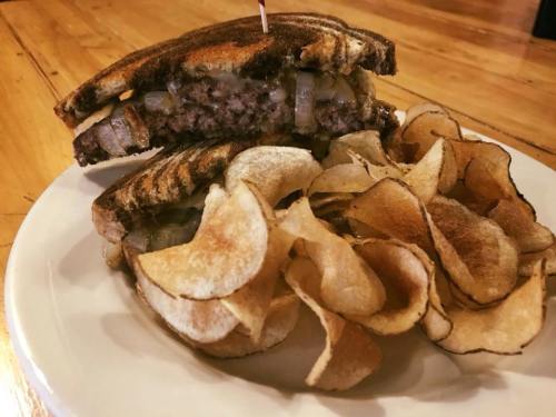 A grilled sandwich with a juicy burger patty and onions, served with a side of crispy potato chips.
