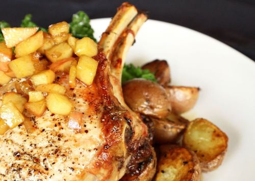 A juicy pork chop topped with diced apples, served with roasted potatoes and greens on a white plate.