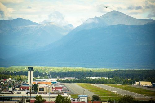 A small plane flies over a mountainous landscape near an airport, with a control tower and runway visible below.