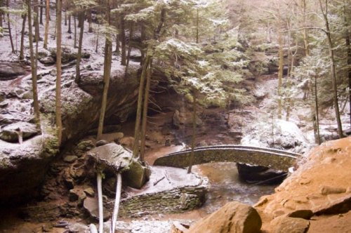 A snow-covered forest scene with a stone bridge over a creek, surrounded by trees and rocky terrain.
