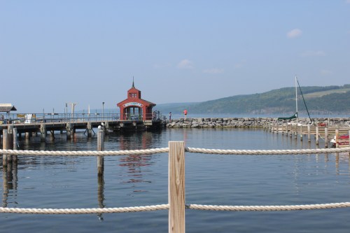 A peaceful lakeside scene featuring a red boathouse, dock, and calm water under a clear blue sky.