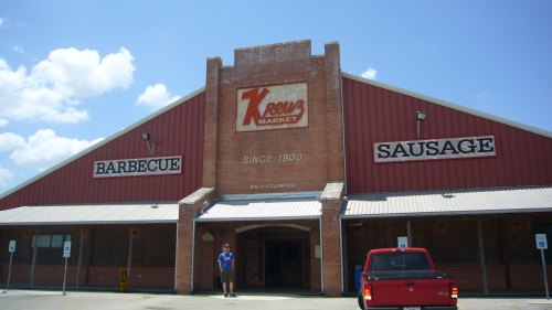 Exterior of Kreuz Market, a brick building with signs for barbecue and sausage, under a clear blue sky.