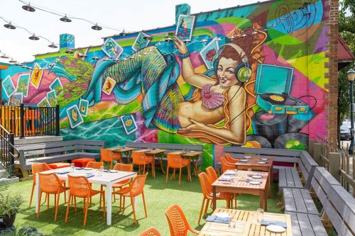 Colorful mural of a mermaid with headphones on a vibrant wall, overlooking a patio with orange chairs and tables.