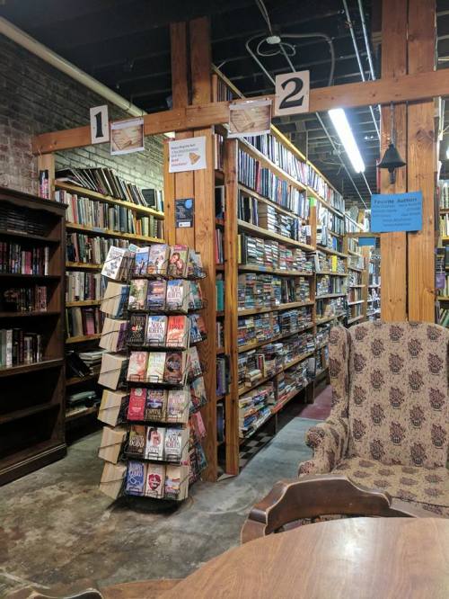 A cozy bookstore aisle with wooden shelves filled with books, a comfy chair, and a display rack of novels.