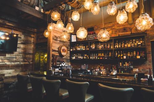 A cozy bar with wooden walls, a well-stocked shelf of liquor, and warm pendant lights hanging above the counter.